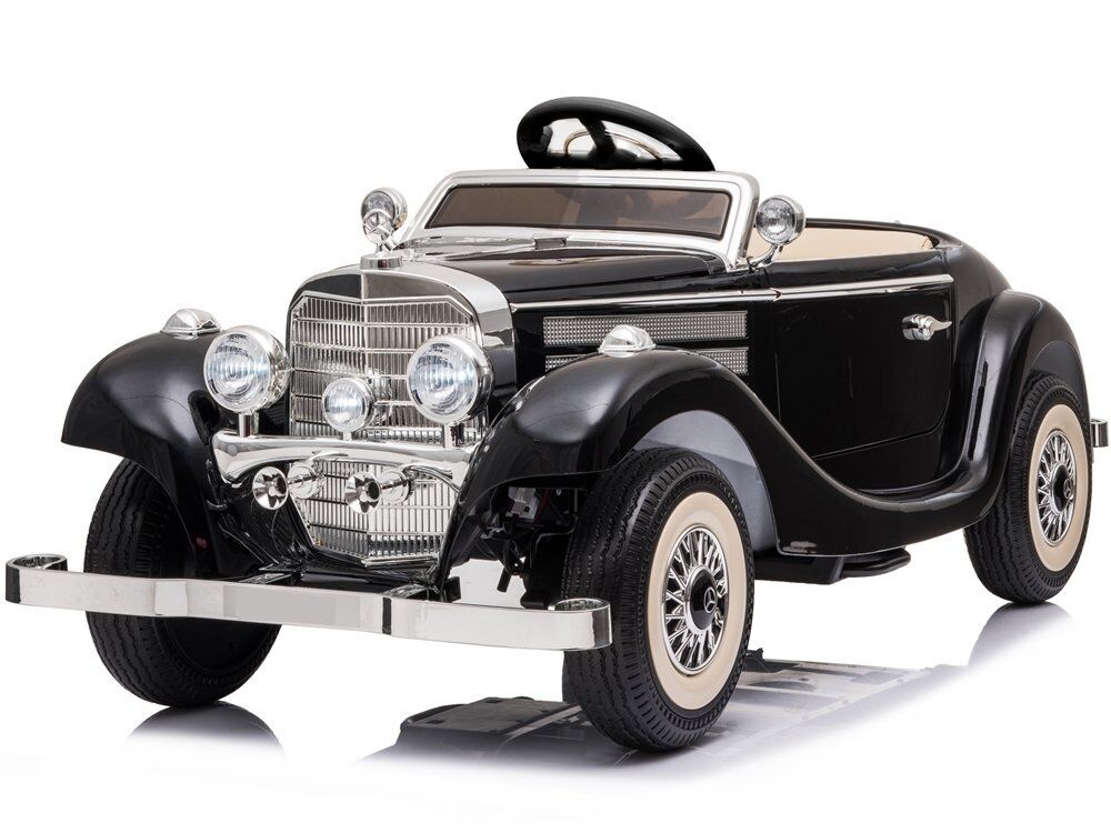Schwarzes, batteriebetriebenes Kinderauto im eleganten Oldtimer-Stil der 1930er. Mit großem Chrom-Kühlergrill, vier Scheinwerfern, Chromstoßstangen und cremefarbenen Weißwandreifen mit Speichenfelgen. Cabrio, vorne links.