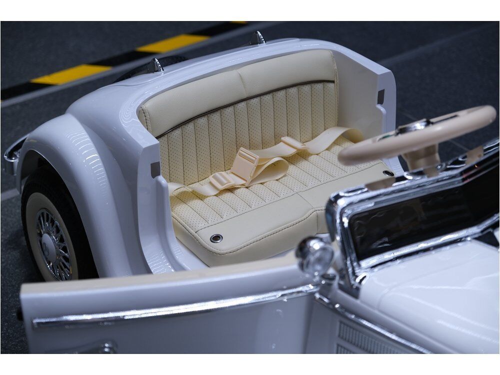 Weißes Kinder-Cabrio im Vintage-Stil mit cremefarbener, perforierter Sitzbank und Sicherheitsgurt. Glänzende Chromdetails, weißes Lenkrad, klassische Radkästen mit Weißwandreifen. Hintergrund: dunkle Fahrbahn mit gelben Markierungen.