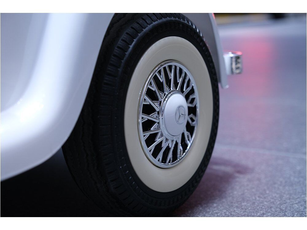 Nahaufnahme des hinteren Reifens eines klassischen weißen Mercedes-Benz. Breite cremefarbene Seitenwand, chromglänzende Radkappe mit Drahtspeichen und Mercedes-Stern in der Mitte. Boden im Hintergrund leicht unscharf.