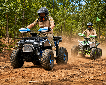 Két, védőruházatban lévő motoros vezeti quadjait egy poros erdei úton, miközben az elöl haladó jármű, egy fekete-kék quad, porfelhőt kavar. A háttérben egy második motoros követi őket egy zöld-fehér quaddal, sűrű fák között.
